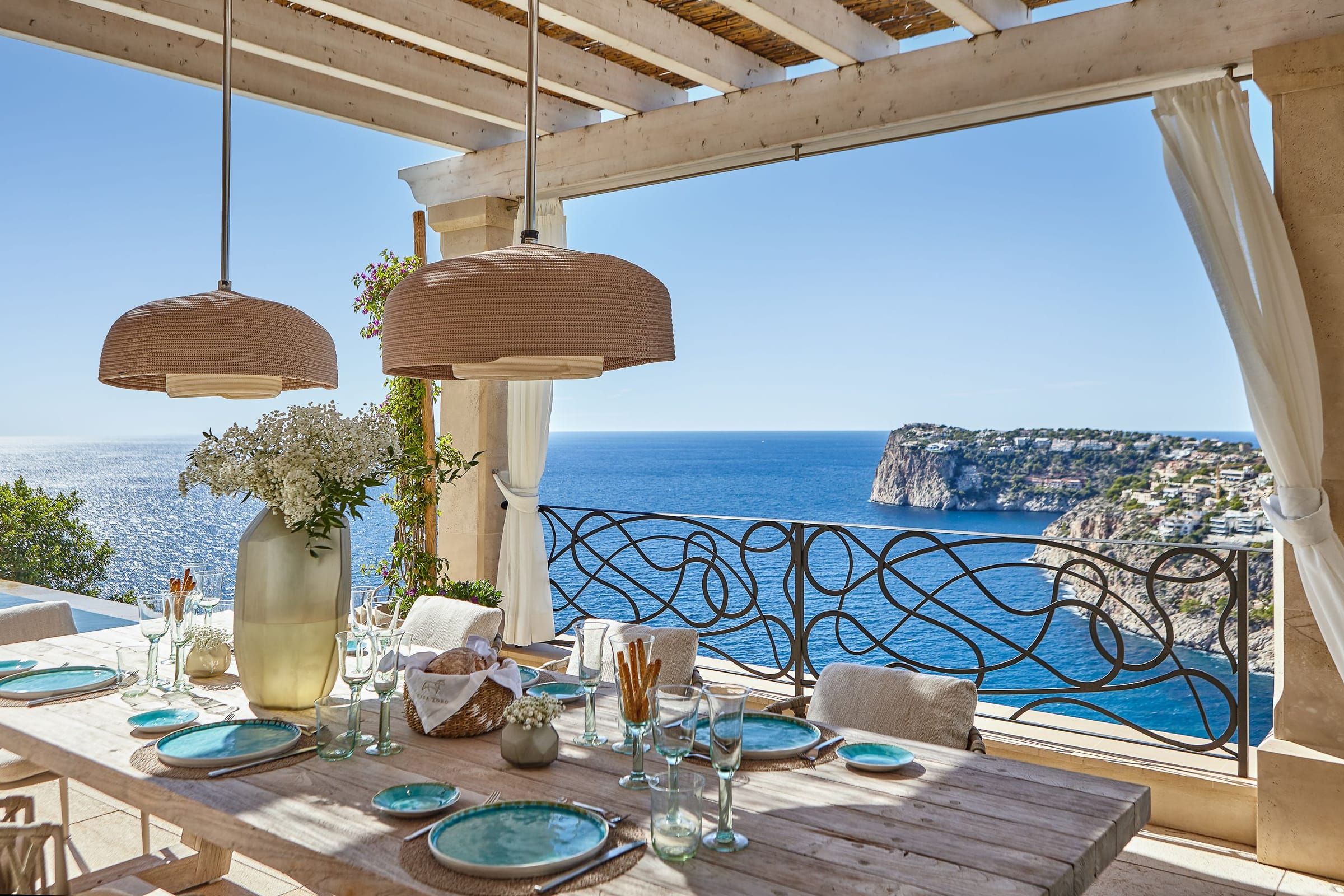 Essbereich im Freien unter der Pergola mit geraffter weißer Gardine und Blick auf Meer und Steilküste