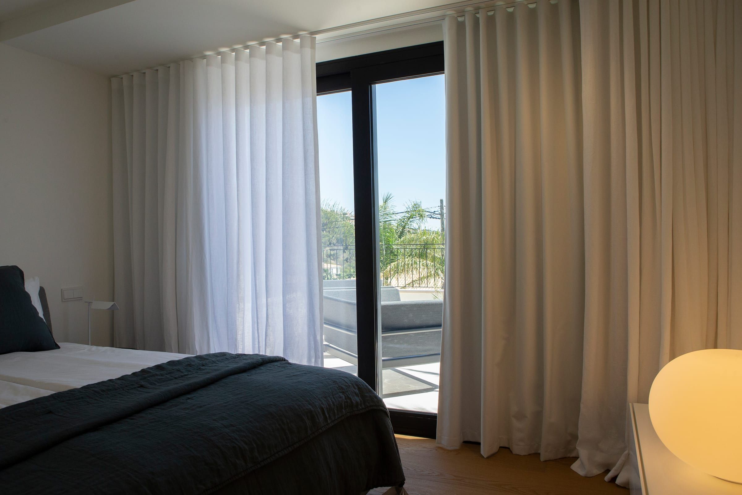 Schlafzimmer mit weißem Store und beigefarbenem Wellenvorhang, zur Terrasse hin geöffnet
