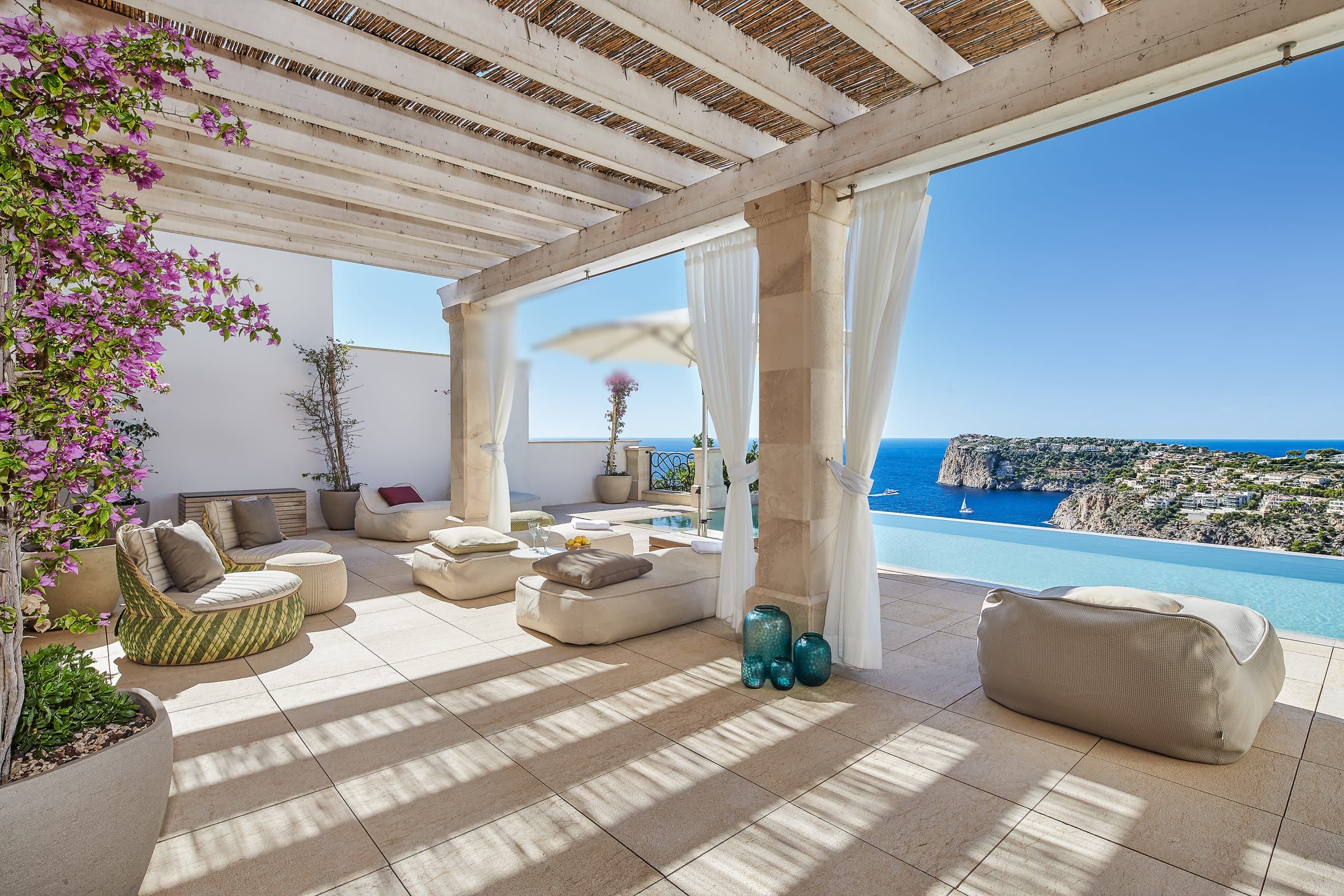 Lounge-Bereich mit Außenvorhängen zwischen den Säulen, Infinity-Pool und Meer im Hintergrund