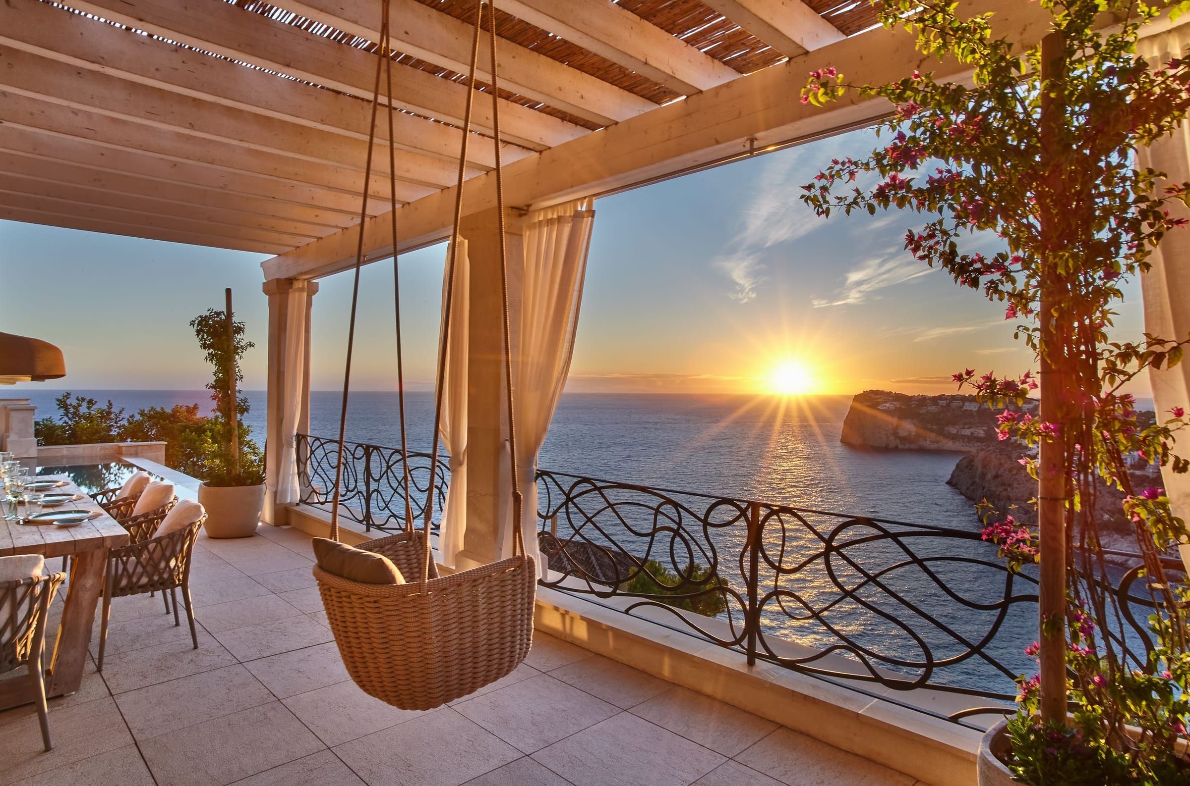 Terrasse mit Hängesessel und weißen Vorhängen an der Pergola bei Sonnenuntergang