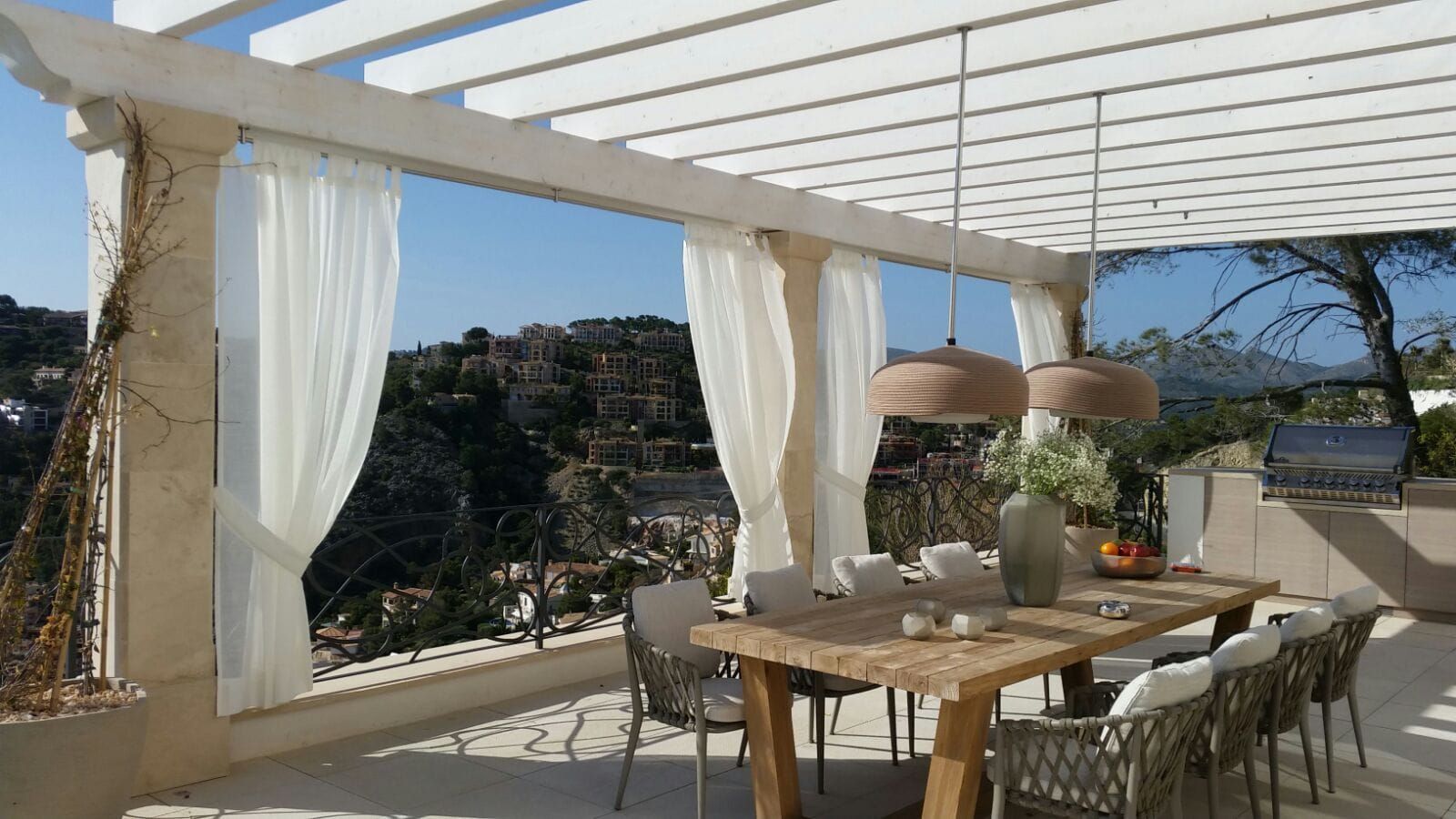 Esstisch im Freien unter einer weißen Pergola mit gerafften Vorhängen und Blick auf die Hügel